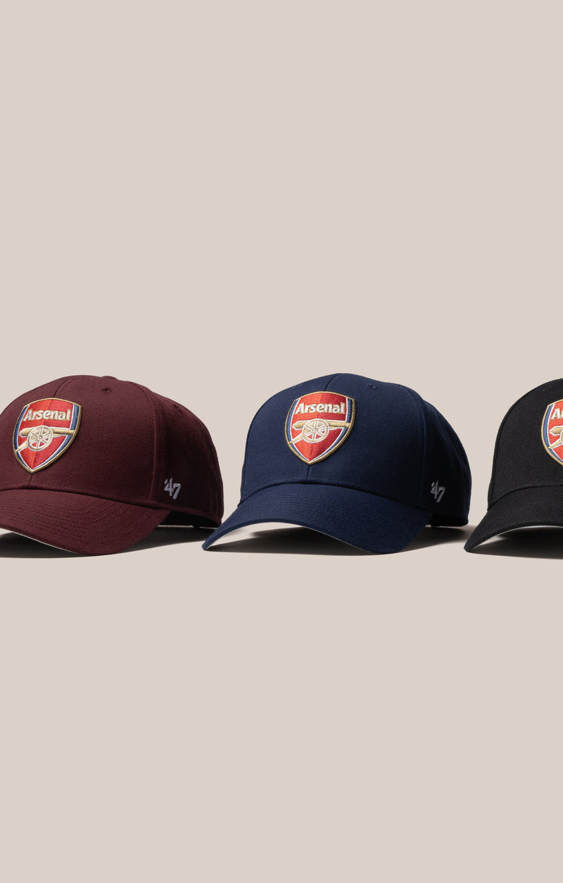 Three Arsenal baseball caps in maroon, navy blue, and black, lined up horizontally against a solid beige background. Each cap features the Arsenal club crest on the front and a small white '47 logo on the side.