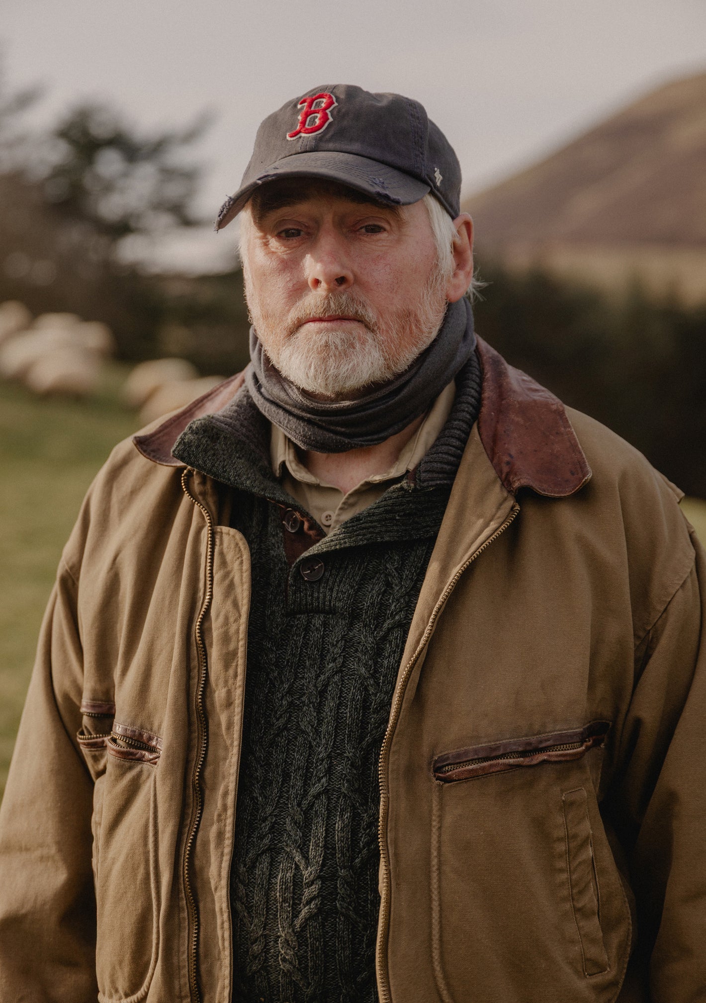 A picture of an older man in a barn jacket wearing a navy '47 Boston Red Sox hat with mountains in the background 